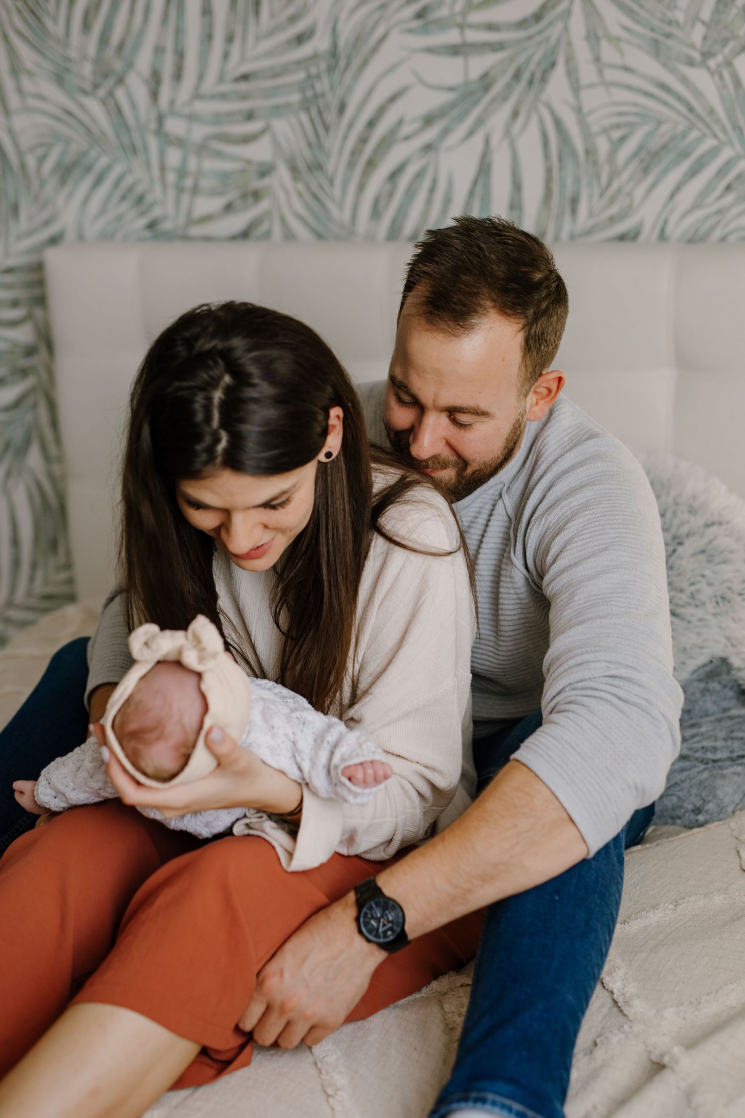 Parents sharing tender moment with newborn against palm leaf wallpaper.