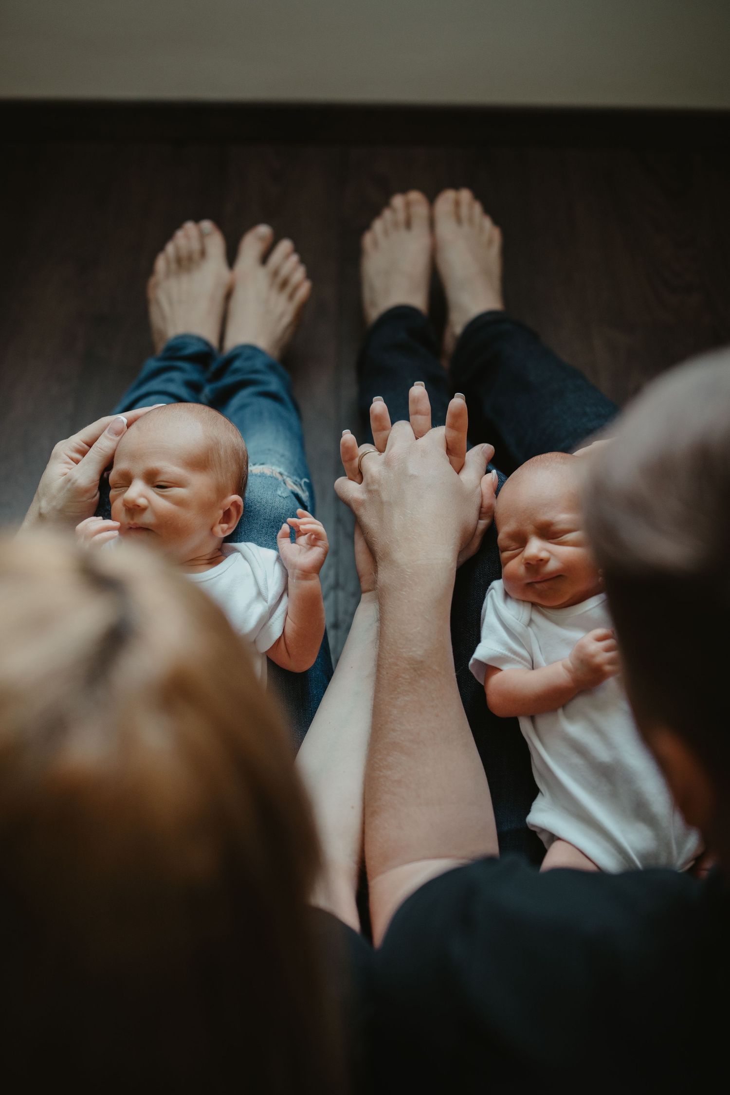 Two babies lying on wooden floor while parents' hands reach toward them.