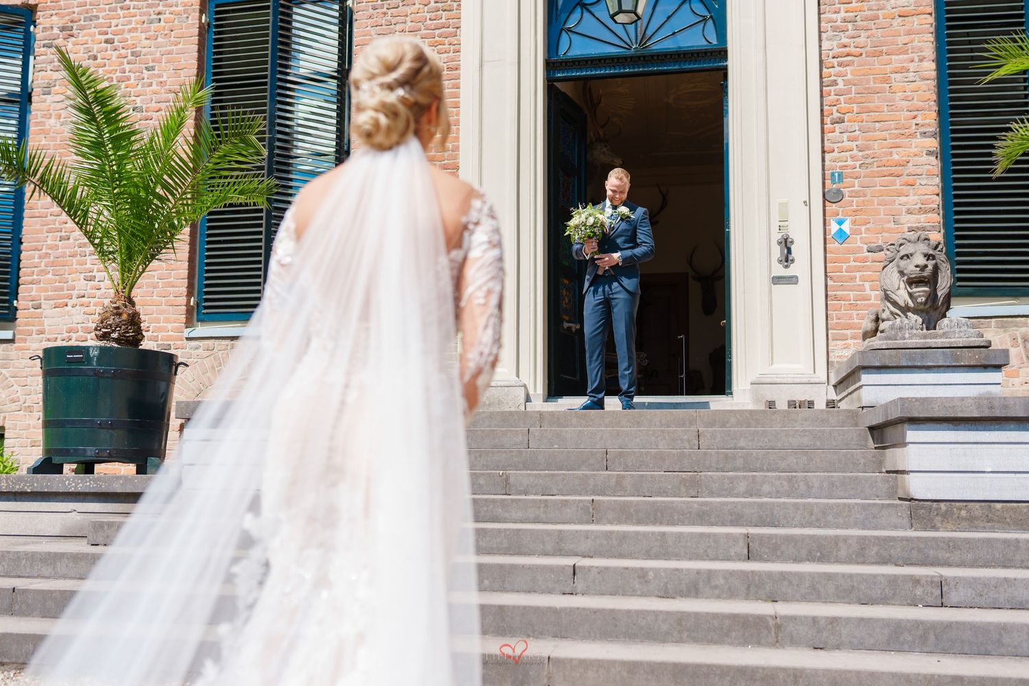 A bride in a flowing veil ascends stone steps where someone stands in a doorway taking a photo.