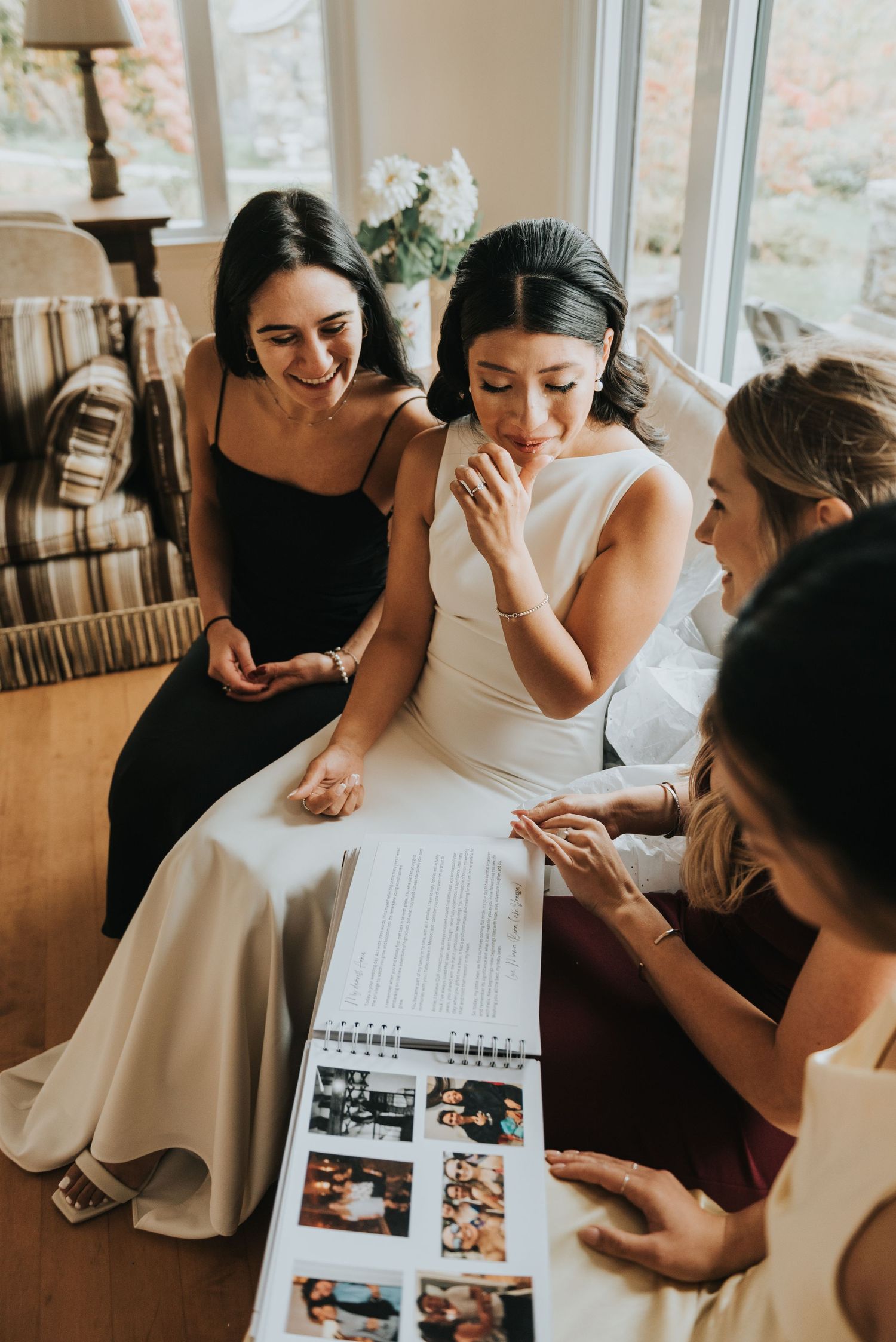 Women in formal attire gather on a couch looking at a photo album together in a sunlit room.