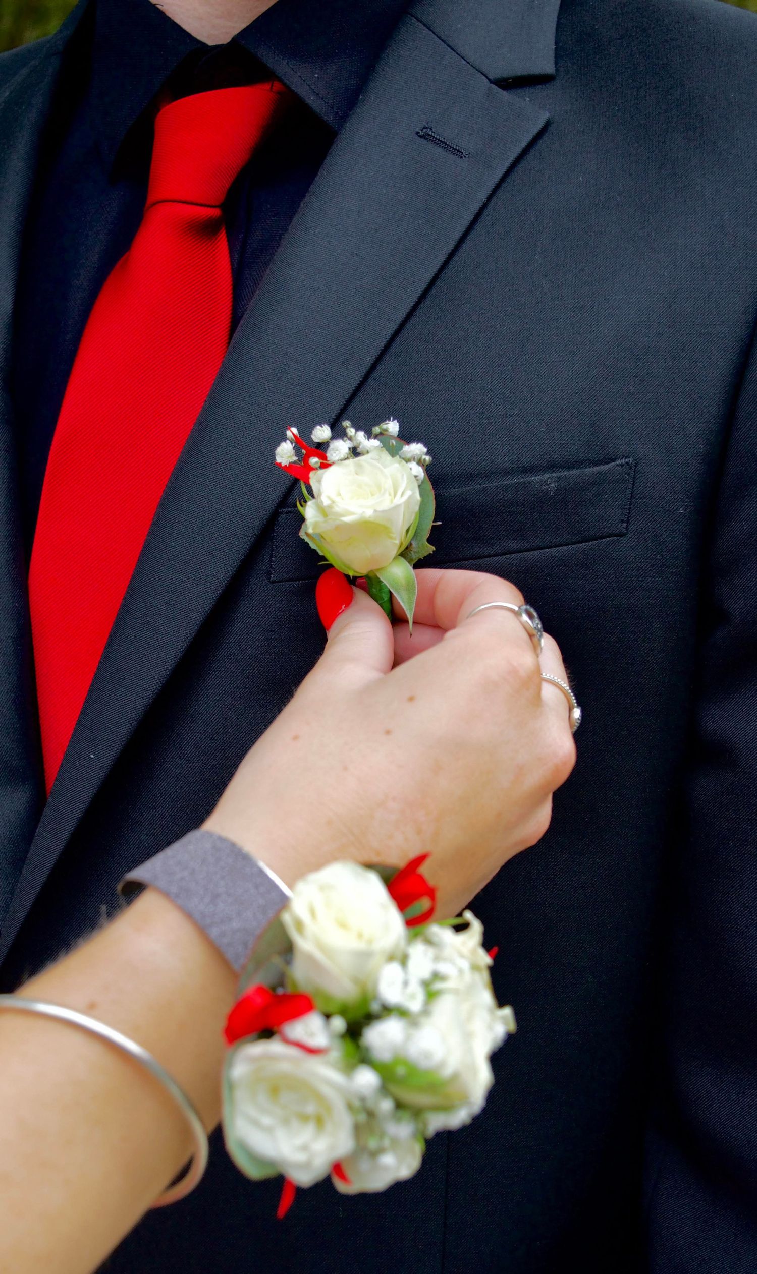Hands pinning a white rose boutonniere on a dark suit jacket with red tie.