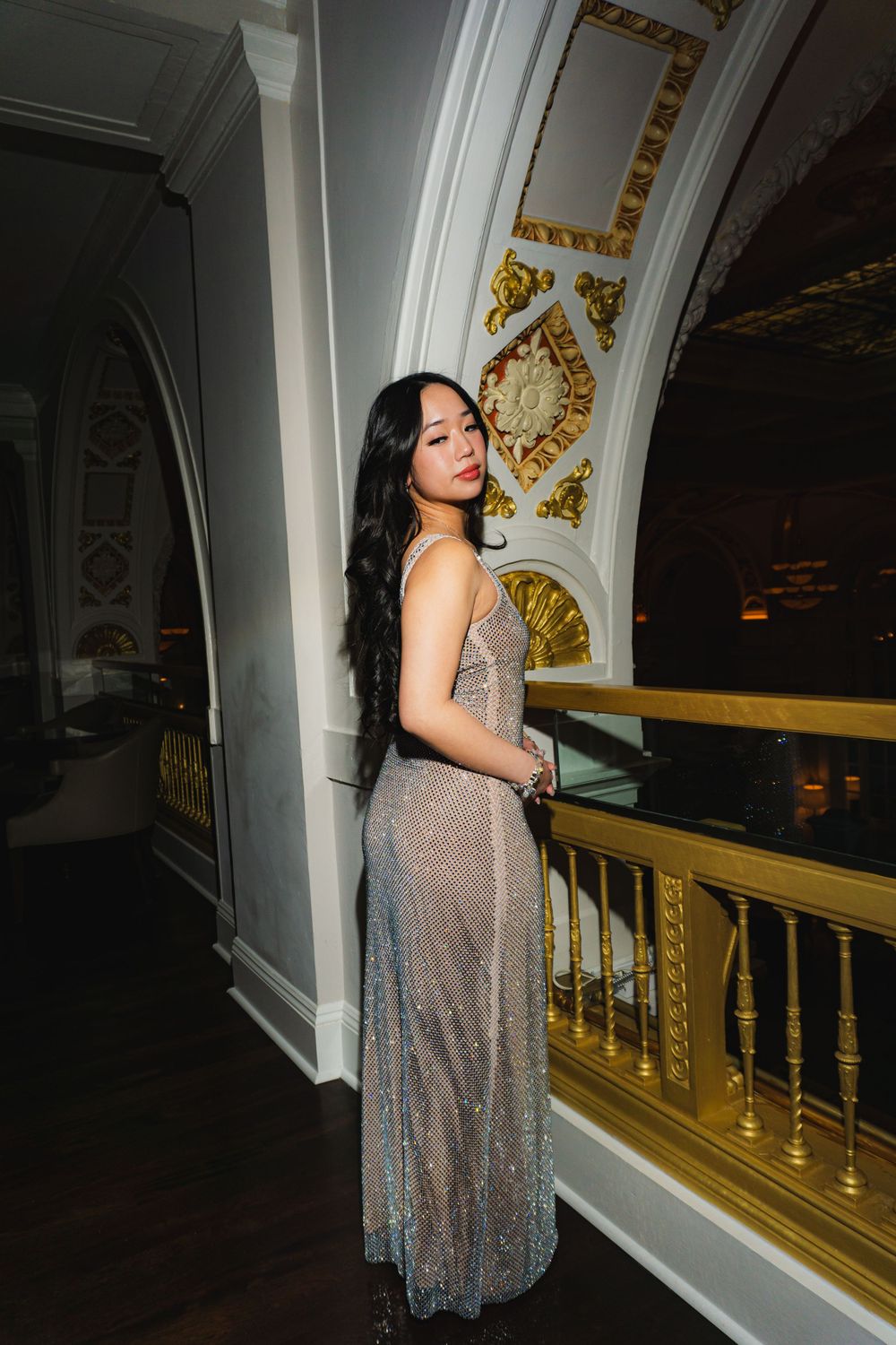 Figure in shimmering evening gown standing by decorative balcony.