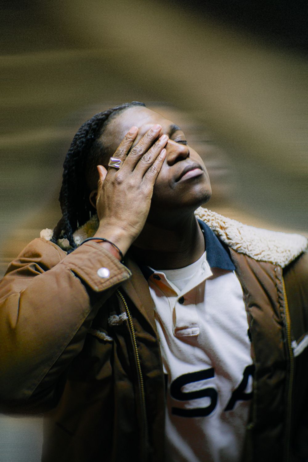 Person in brown jacket and USA shirt covers face with hand in artistic portrait.