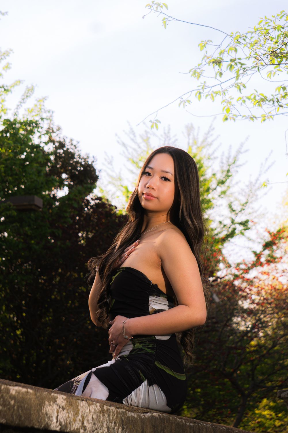 A person in a black dress poses on a stone wall during sunset with trees in the background.