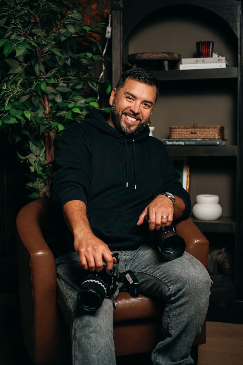 Smiling man in black hoodie sitting in leather chair holding camera, with bookshelf and green plant in background.