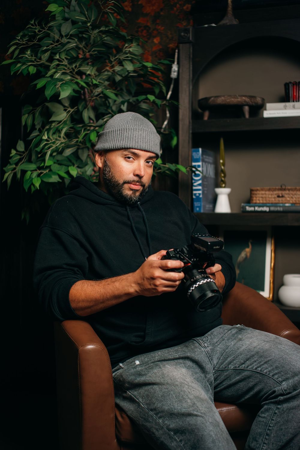 Man in gray beanie and black hoodie sitting in leather chair holding a DSLR camera indoors.