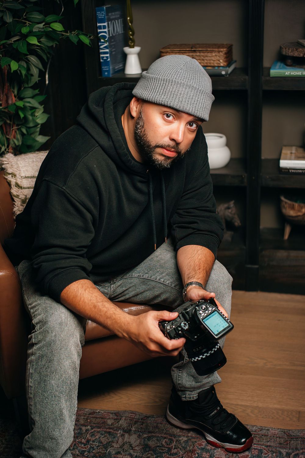 Bearded man in gray beanie and black hoodie sits holding a DSLR camera indoors near bookshelves and plants.