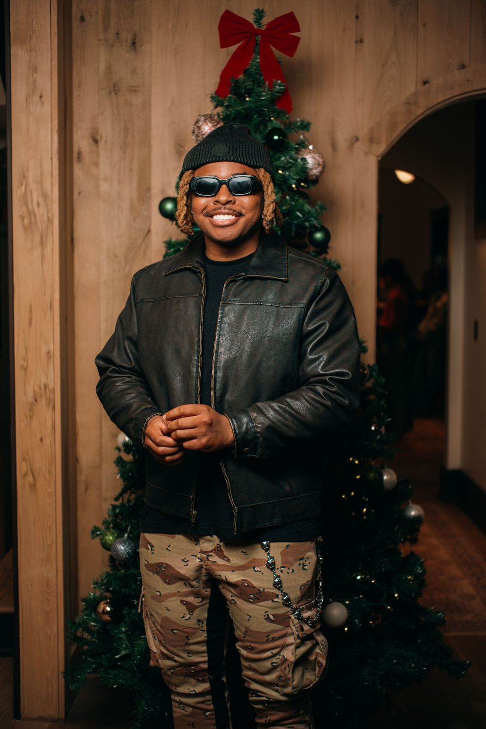 Smiling man in sunglasses and leather jacket poses in front of decorated Christmas tree indoors.