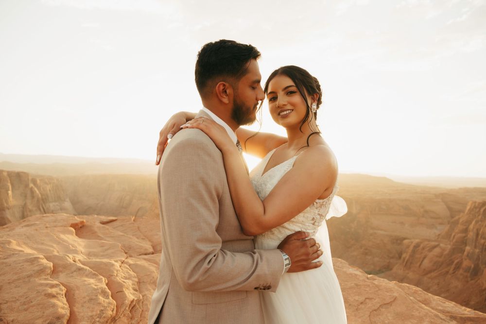 A couple in wedding attire embraces while standing at the edge of a scenic canyon at sunset.