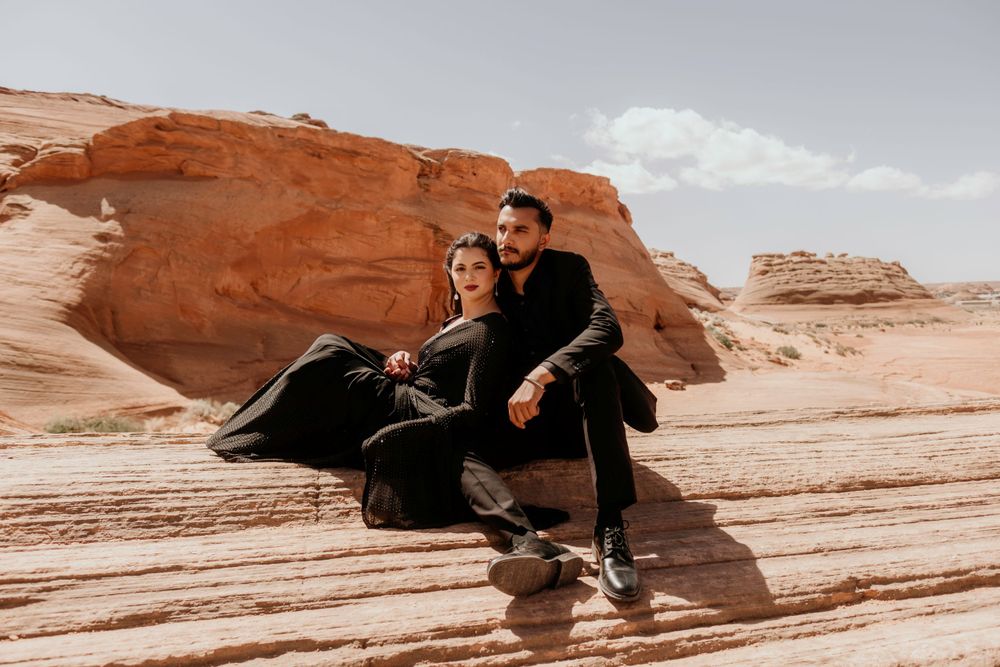 Two people wearing black clothing sit together on red sandstone rock formations in a desert landscape.