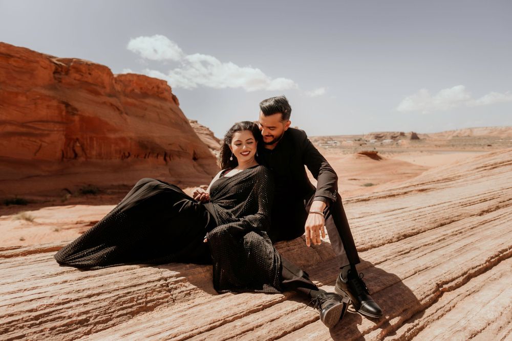 A couple sits together on red sandstone formations in a desert landscape under cloudy skies.