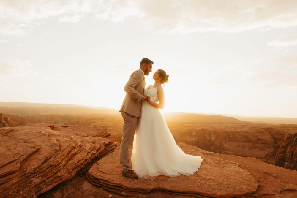 A bridal couple stands silhouetted against a golden sunset on red sandstone cliffs in the desert.