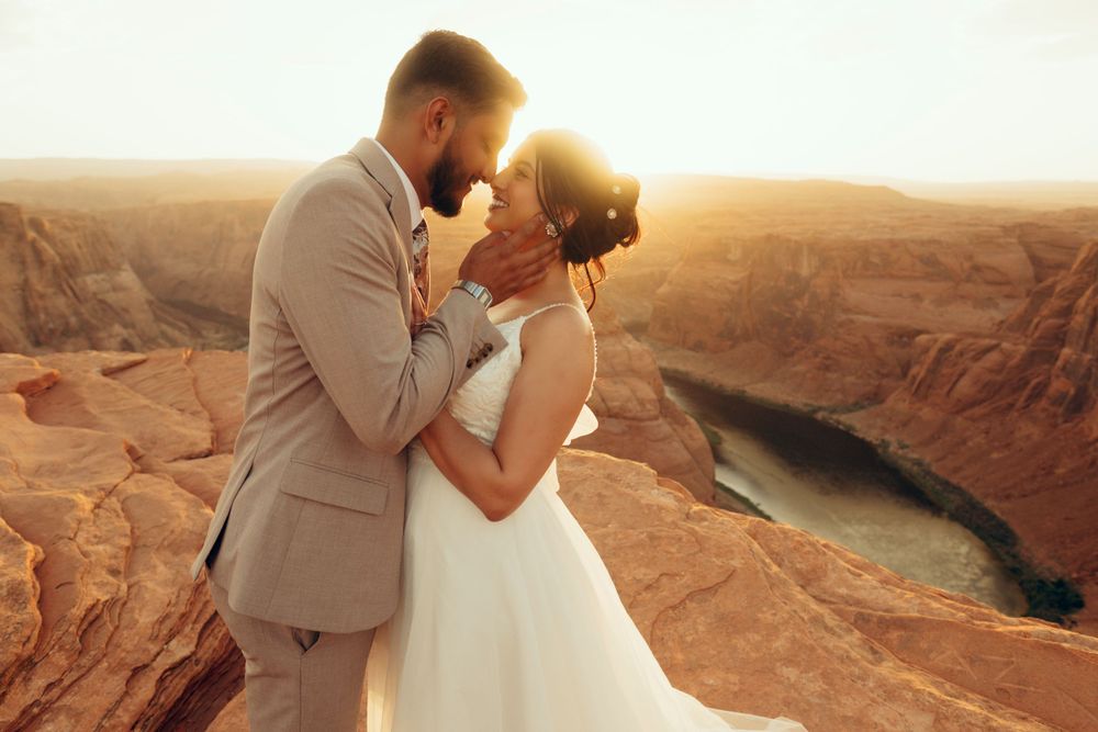 A romantic moment captured between a couple in wedding attire standing atop red rock cliffs overlooking a river at sunset.