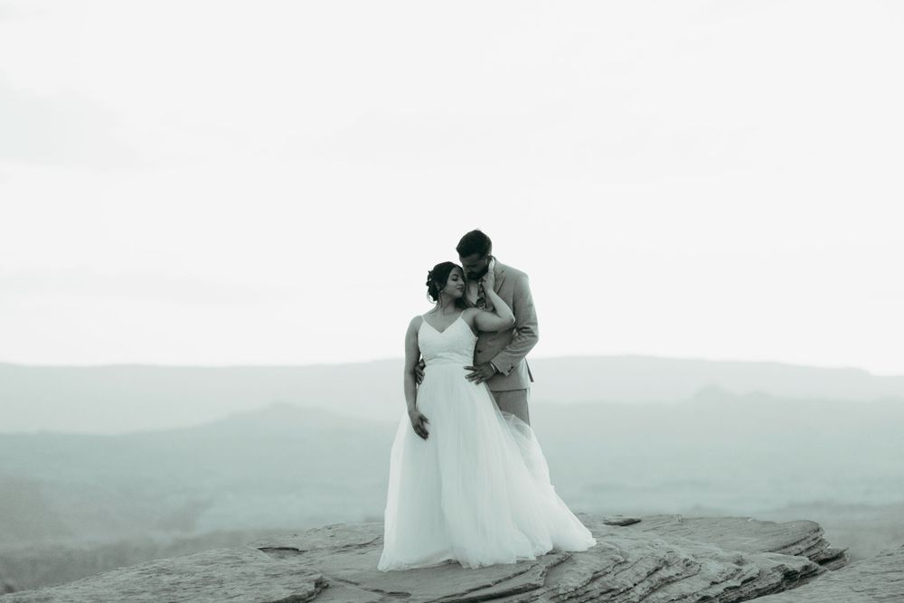 A couple in wedding attire embrace on a cliff edge overlooking a misty mountain landscape in black and white.