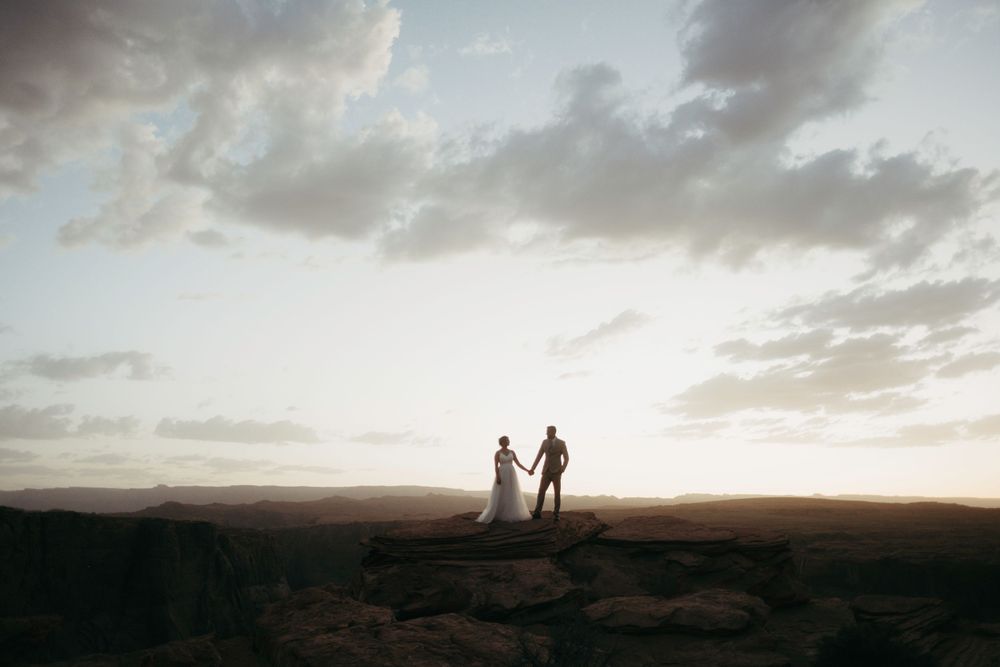 Silhouetted couple holds hands on hilltop against dramatic sunset sky and mountainous landscape.