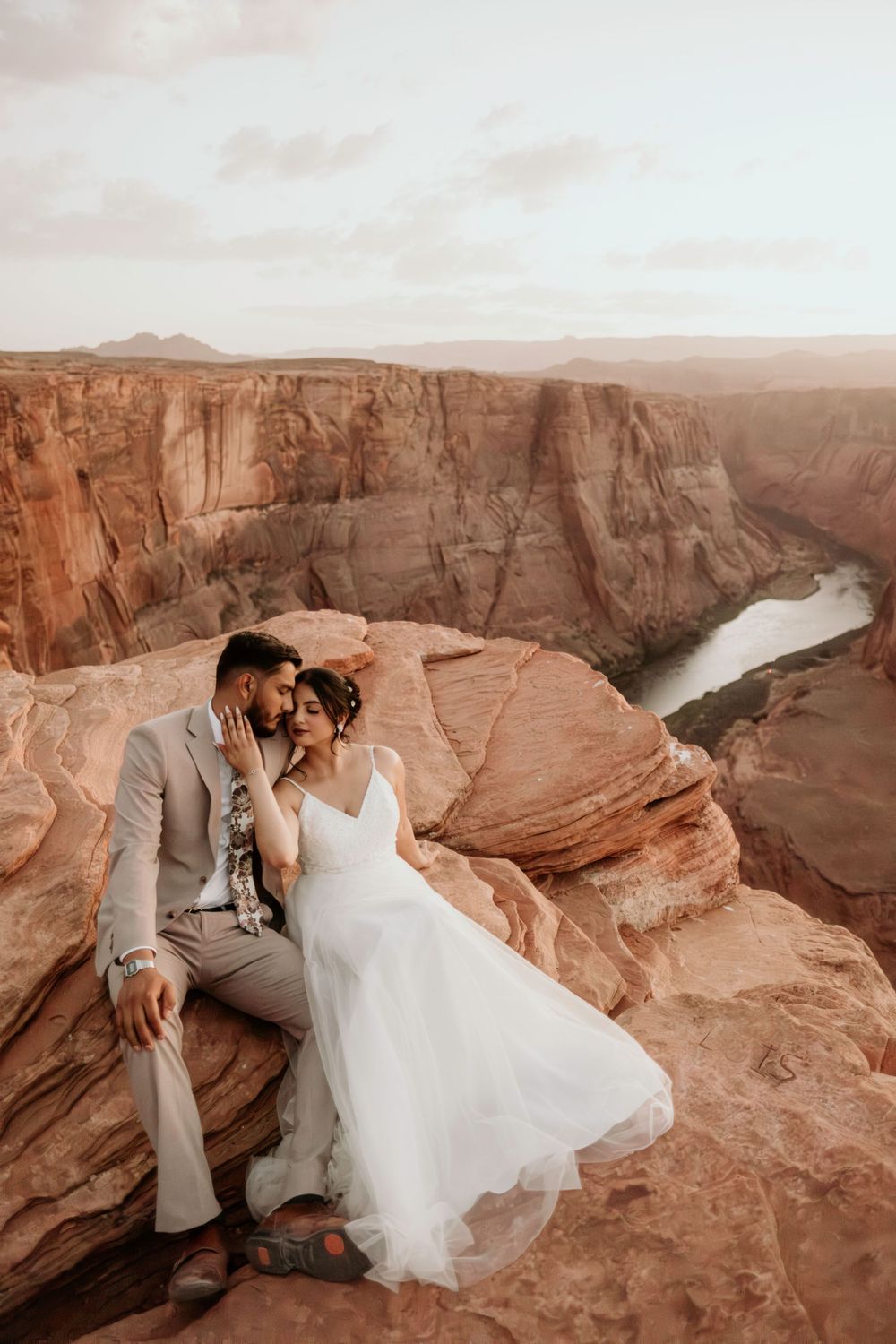 A romantic couple shares an intimate moment on the edge of a majestic canyon with a winding river below at sunset.