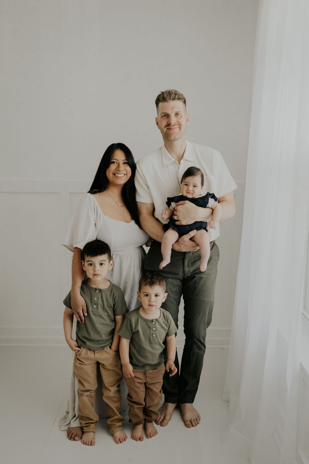 A family of five poses together barefoot against a white wall wearing coordinated neutral-colored outfits.
