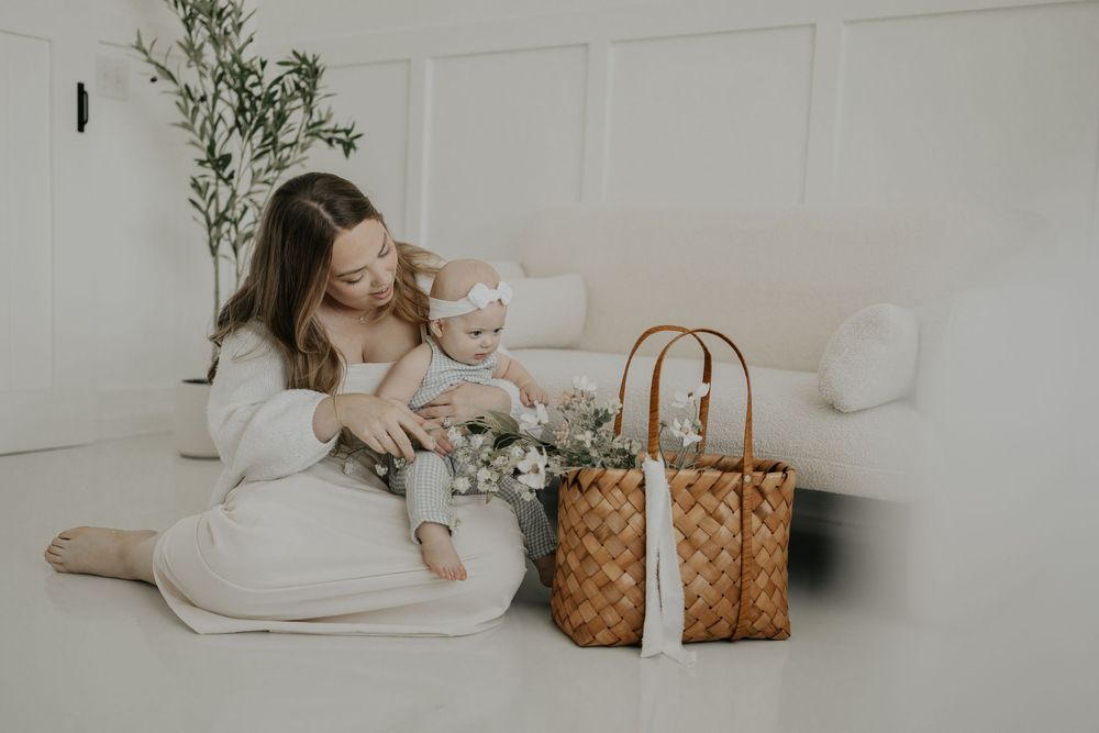 Minimalist white room scene with a woven tan leather tote bag and fresh flower bouquet arrangement on floor.