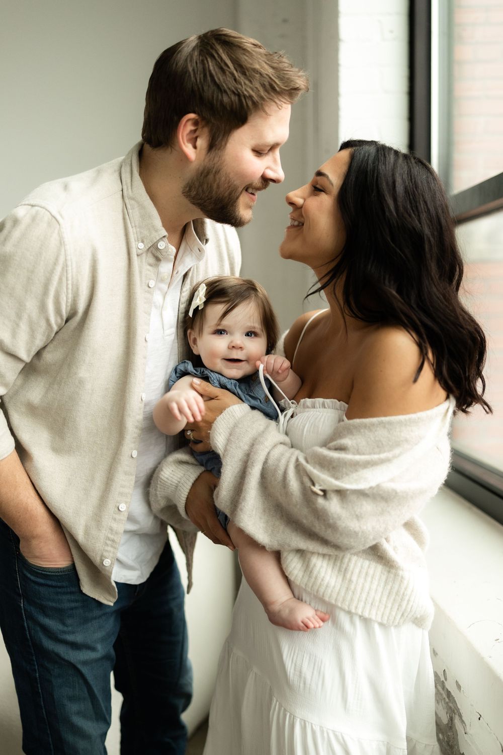 A happy family shares a tender moment together by a bright window, wearing coordinated white and beige outfits.