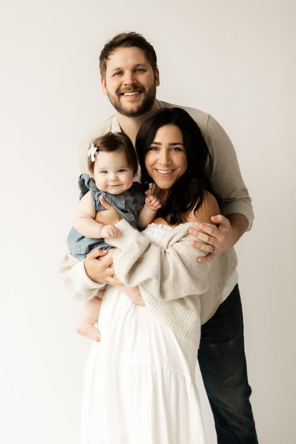 Family portrait on white background with parents embracing their baby in casual attire.