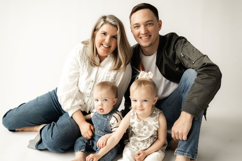 Family of four sitting together on studio floor in casual attire against light background.