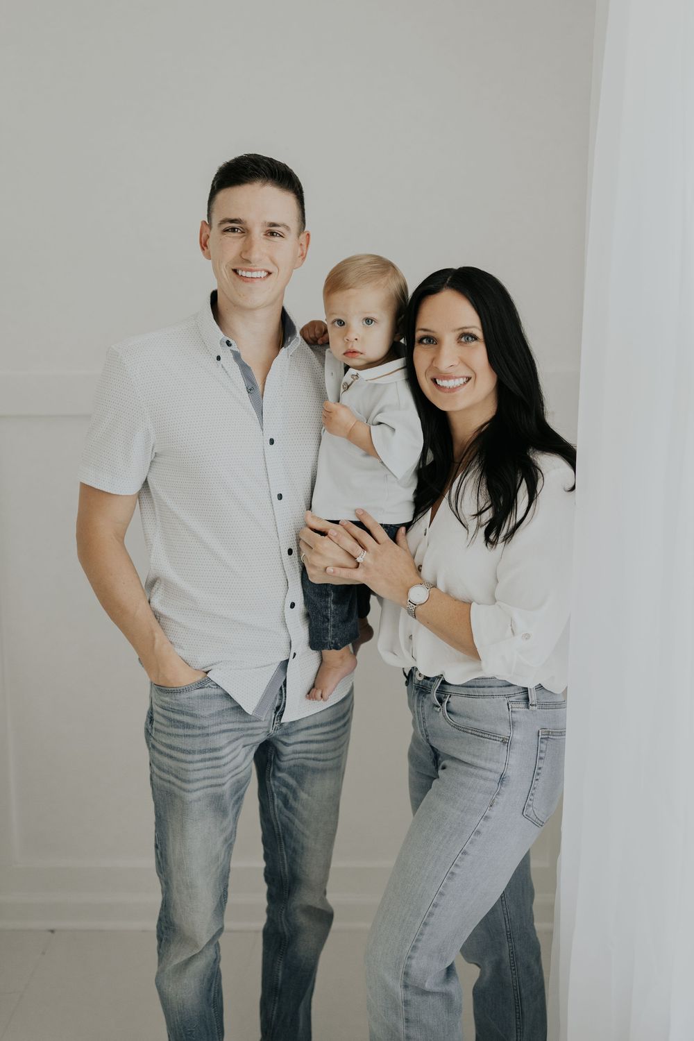 A young family poses together against a white wall wearing coordinated casual outfits in light colors.