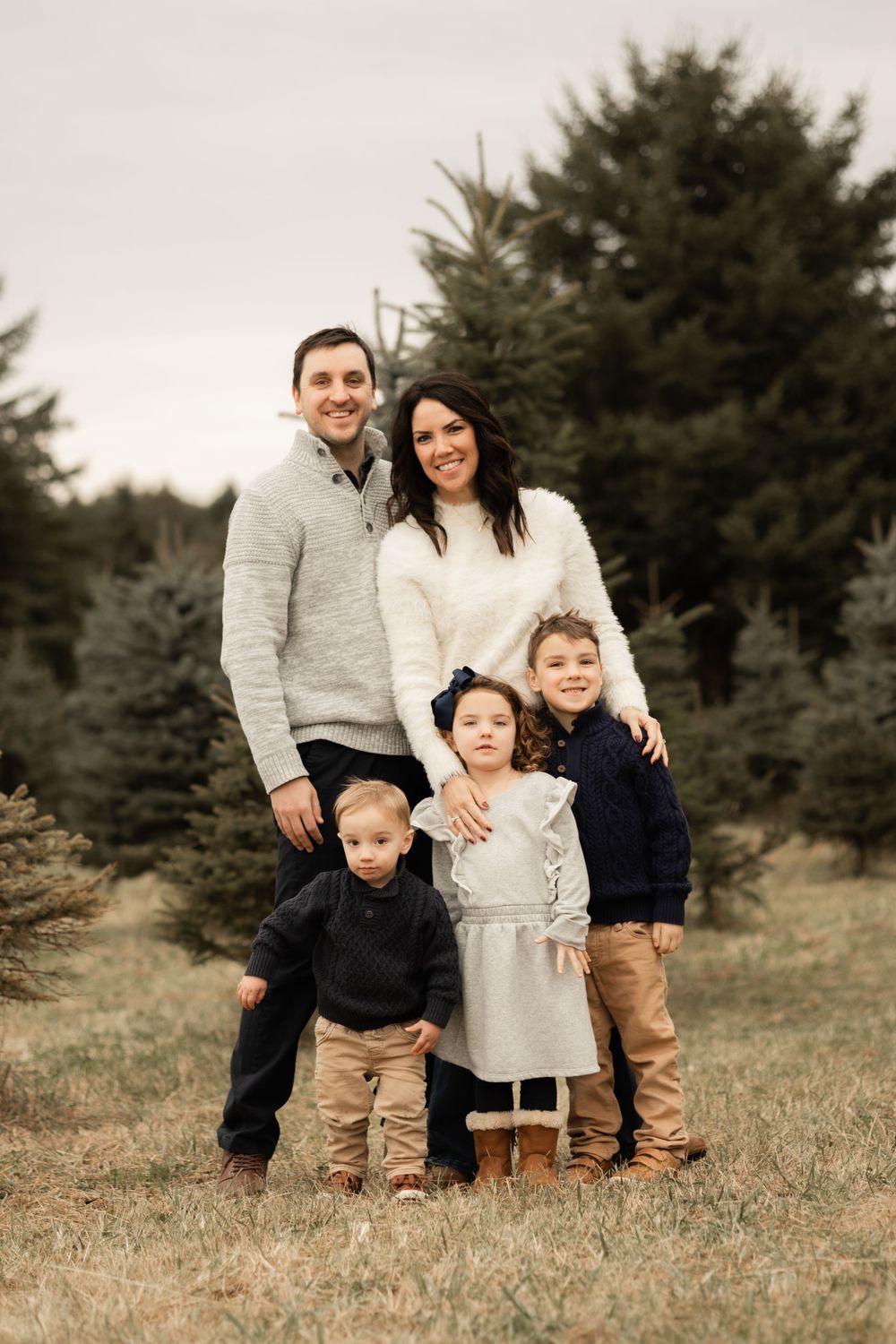 Family portrait outdoors with evergreen trees in background, wearing coordinated casual outfits in neutral colors.