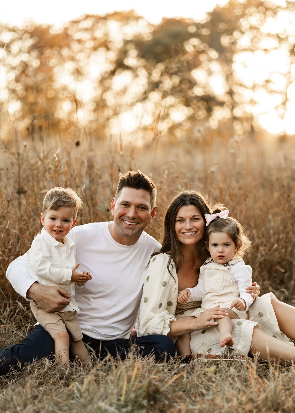 Family sits together in golden hour sunlight among tall grass in a natural outdoor setting.