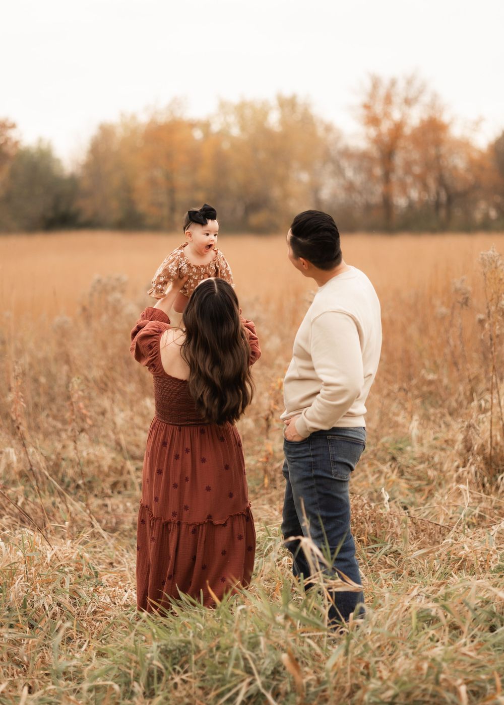 A couple stands together in a golden autumn field during sunset holding their baby in a rustic outdoor setting.