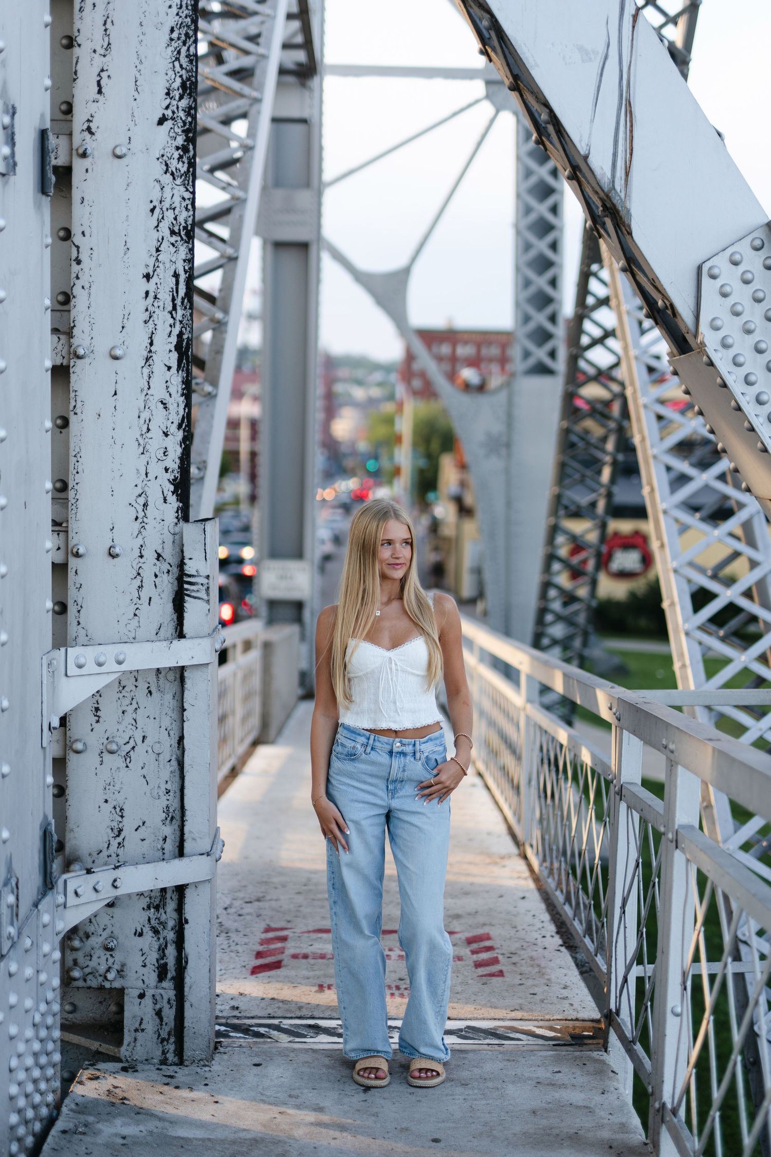Young woman in blue floral sundress poses outdoors near a bridge on a sunny day.