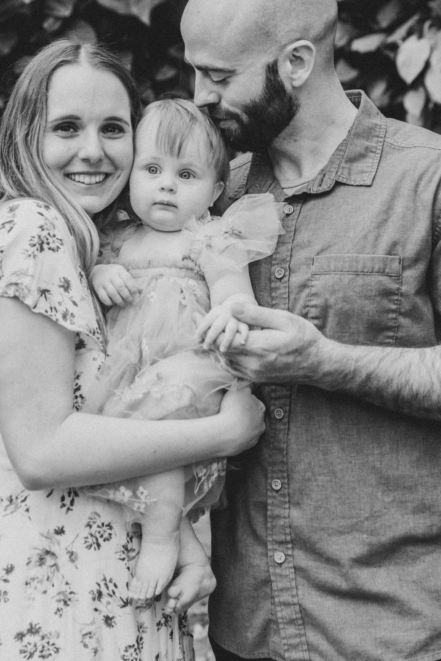 Black and white family portrait showing parents holding their young toddler in a candid outdoor setting.
