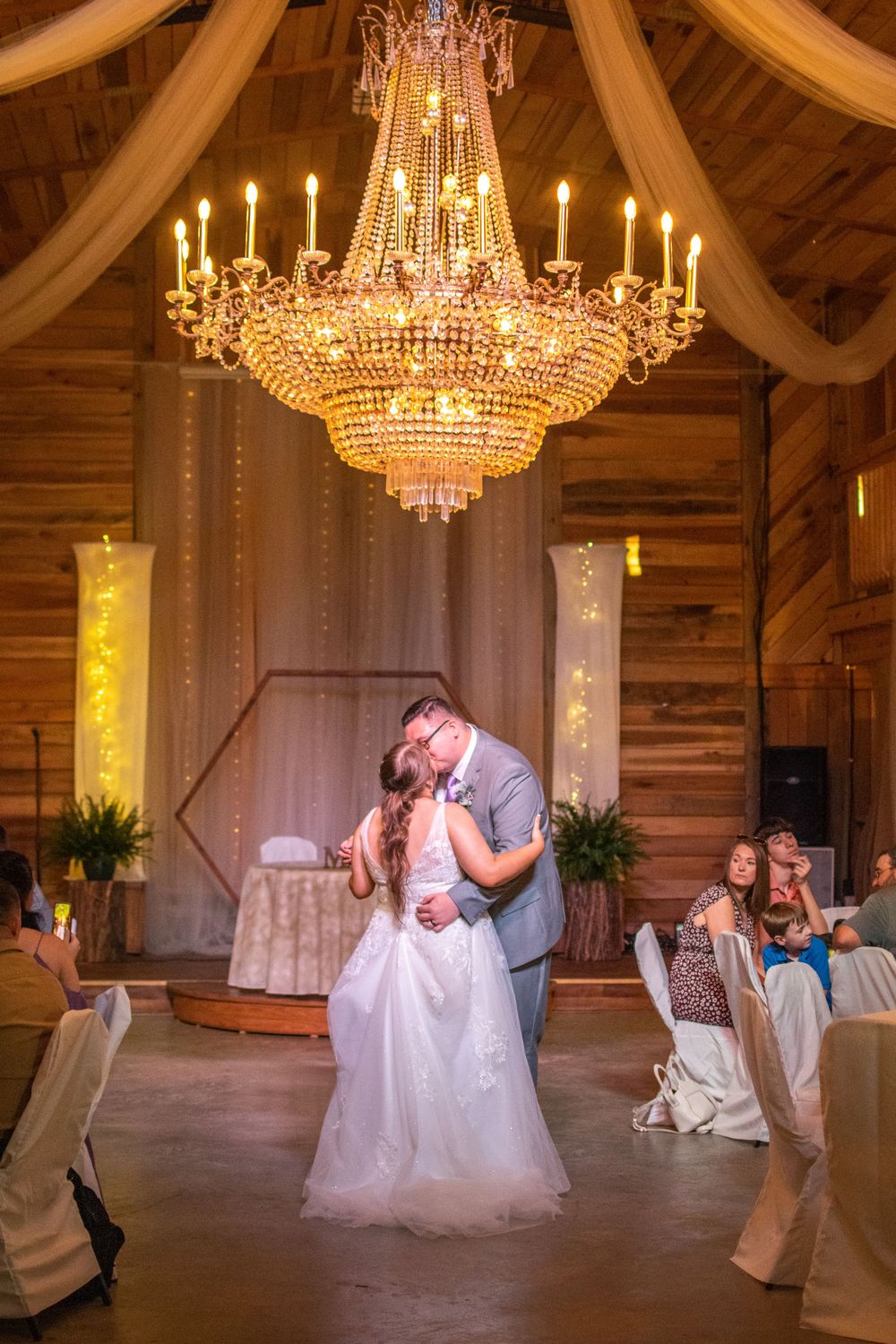 Wedding guests watch as the newlyweds dance beneath crystal chandeliers in an elegant barn venue.