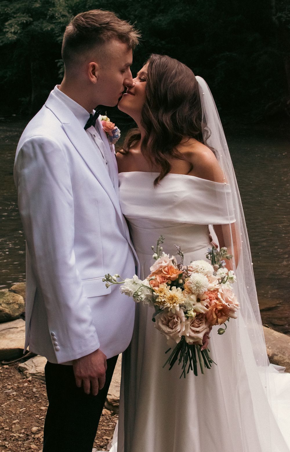 Romantic wedding couple sharing a kiss by a lake with a beautiful floral bouquet.