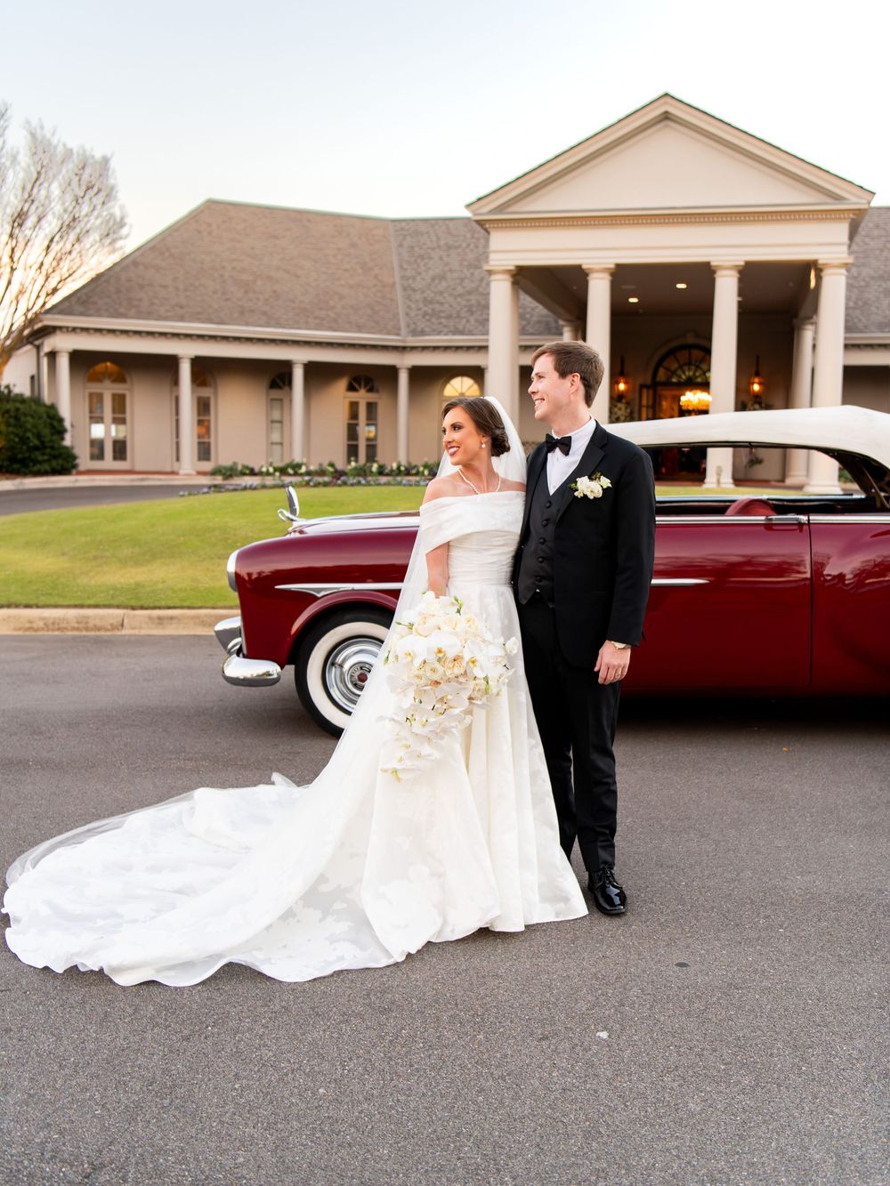 Newlyweds pose beside a vintage red car in front of an elegant wedding venue with columns.