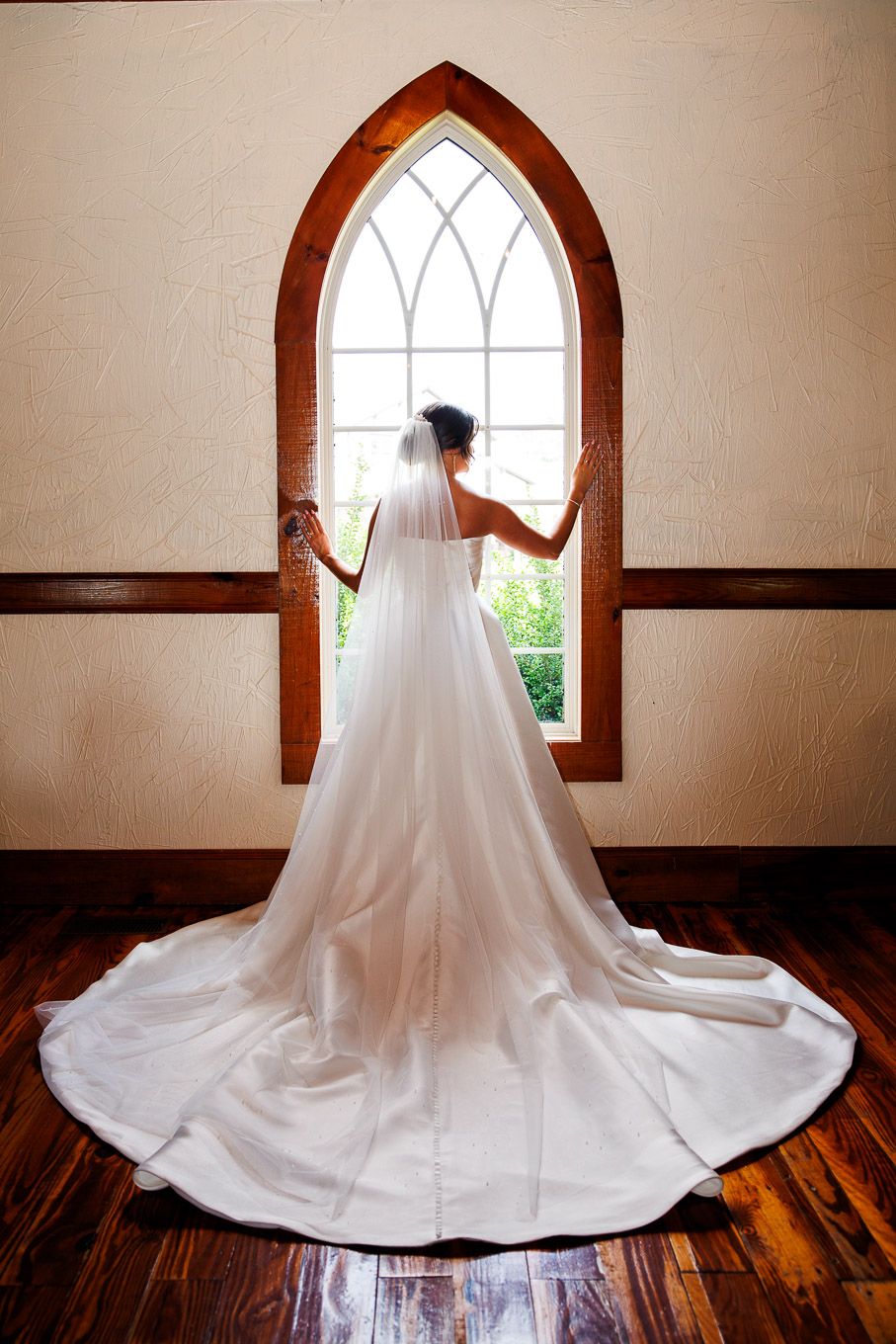 A flowing white wedding dress silhouetted against an arched church window with hardwood floors.