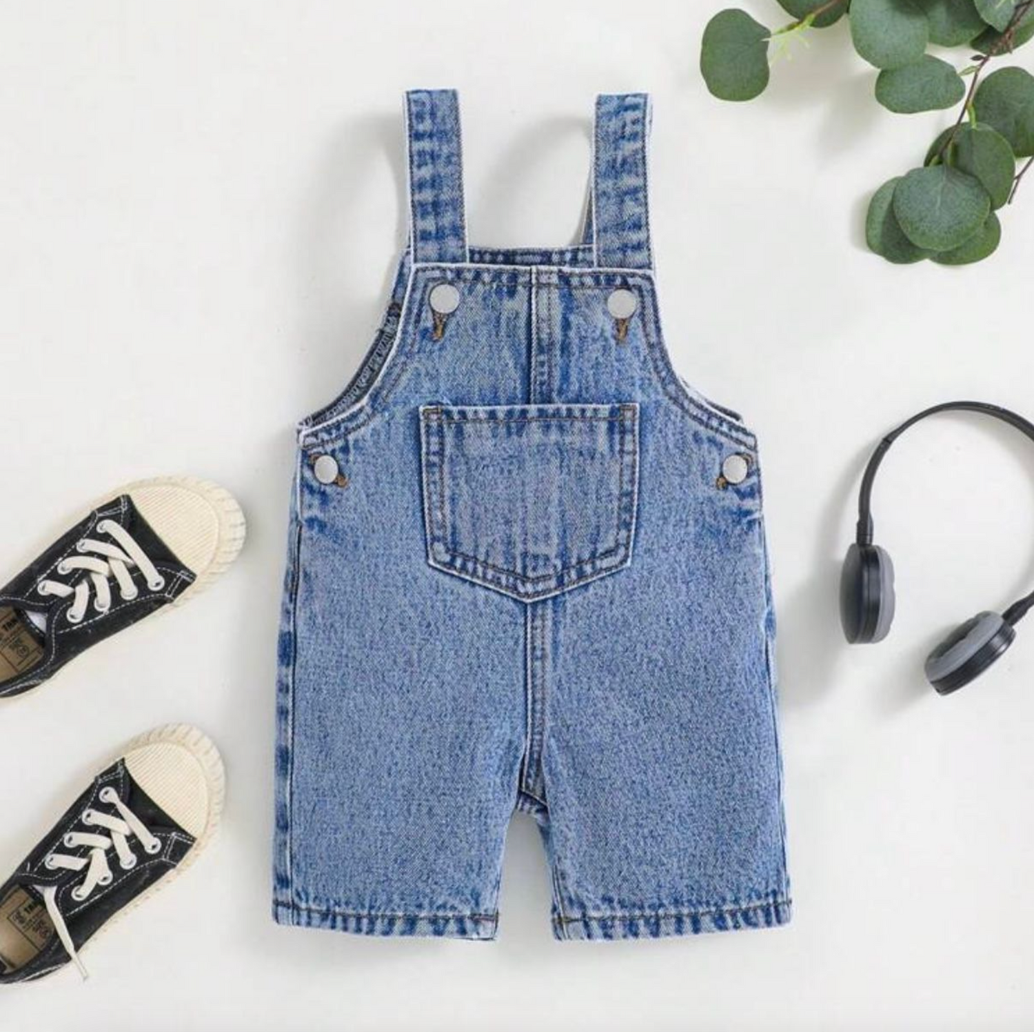 Denim vest with black sneakers and eucalyptus leaves arranged on white background.
