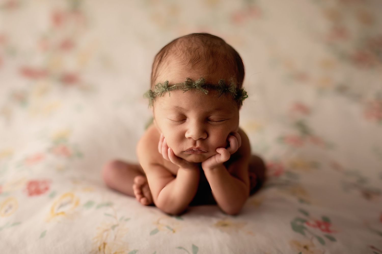 Sleeping newborn lying on floral blanket wearing delicate green leaf headband with hands under chin.