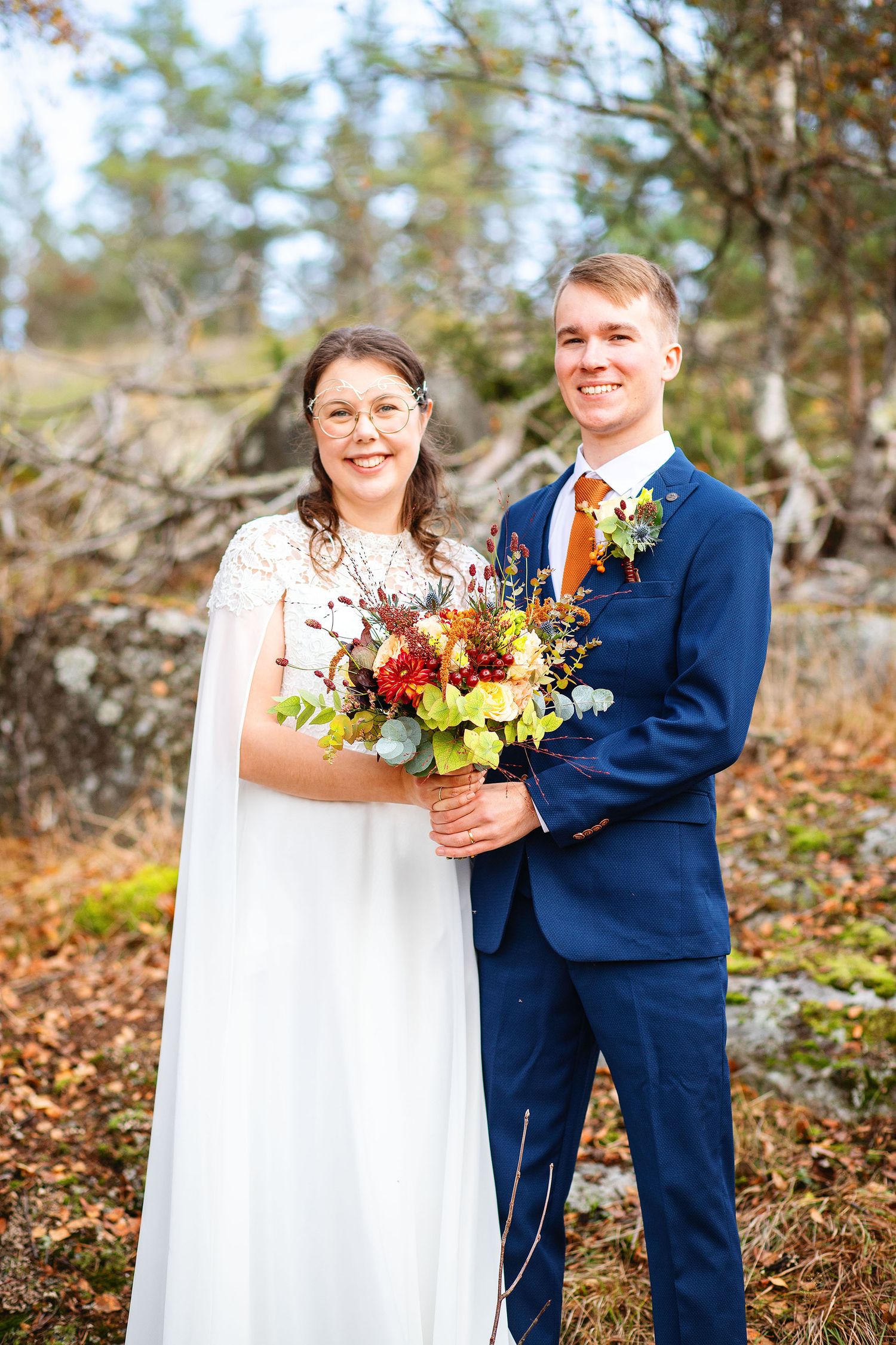 Smiling bride and groom posing outdoors in autumn holding a colorful bouquet, groom wearing a navy suit with orange tie.