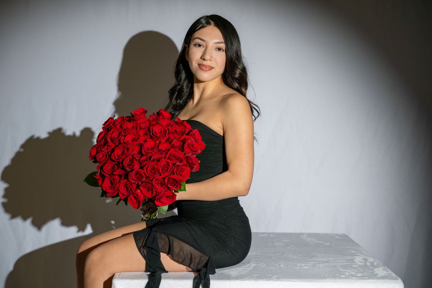 Woman in black dress holding large bouquet of red roses, seated against gray background.