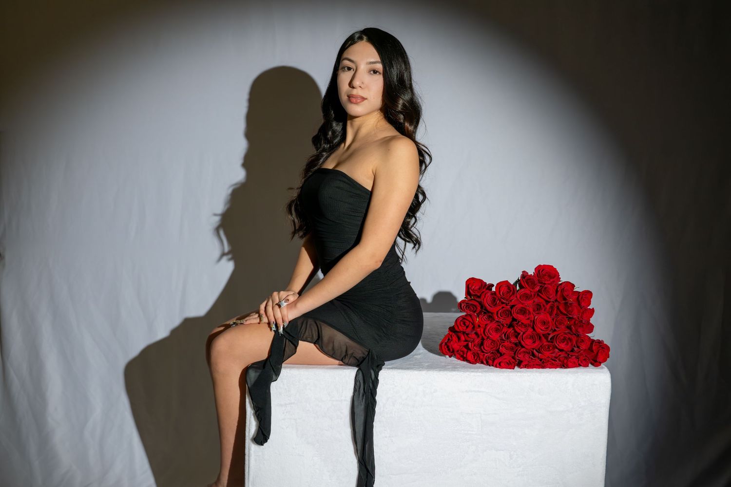 Woman in black dress sits on white cube beside red roses bouquet against grey backdrop.