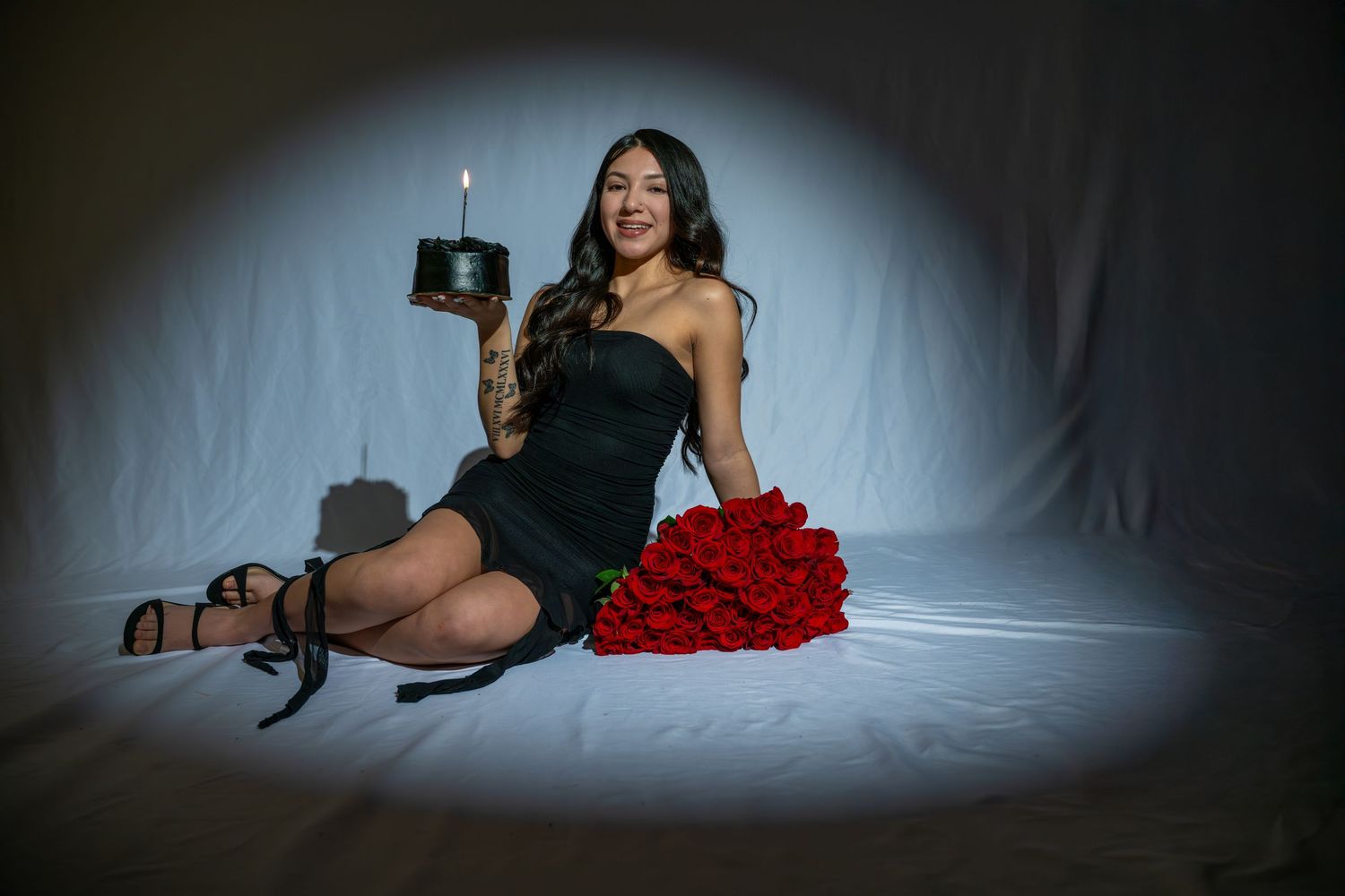 Woman in black dress holding birthday cake with candle, sitting beside red roses bouquet on white backdrop.