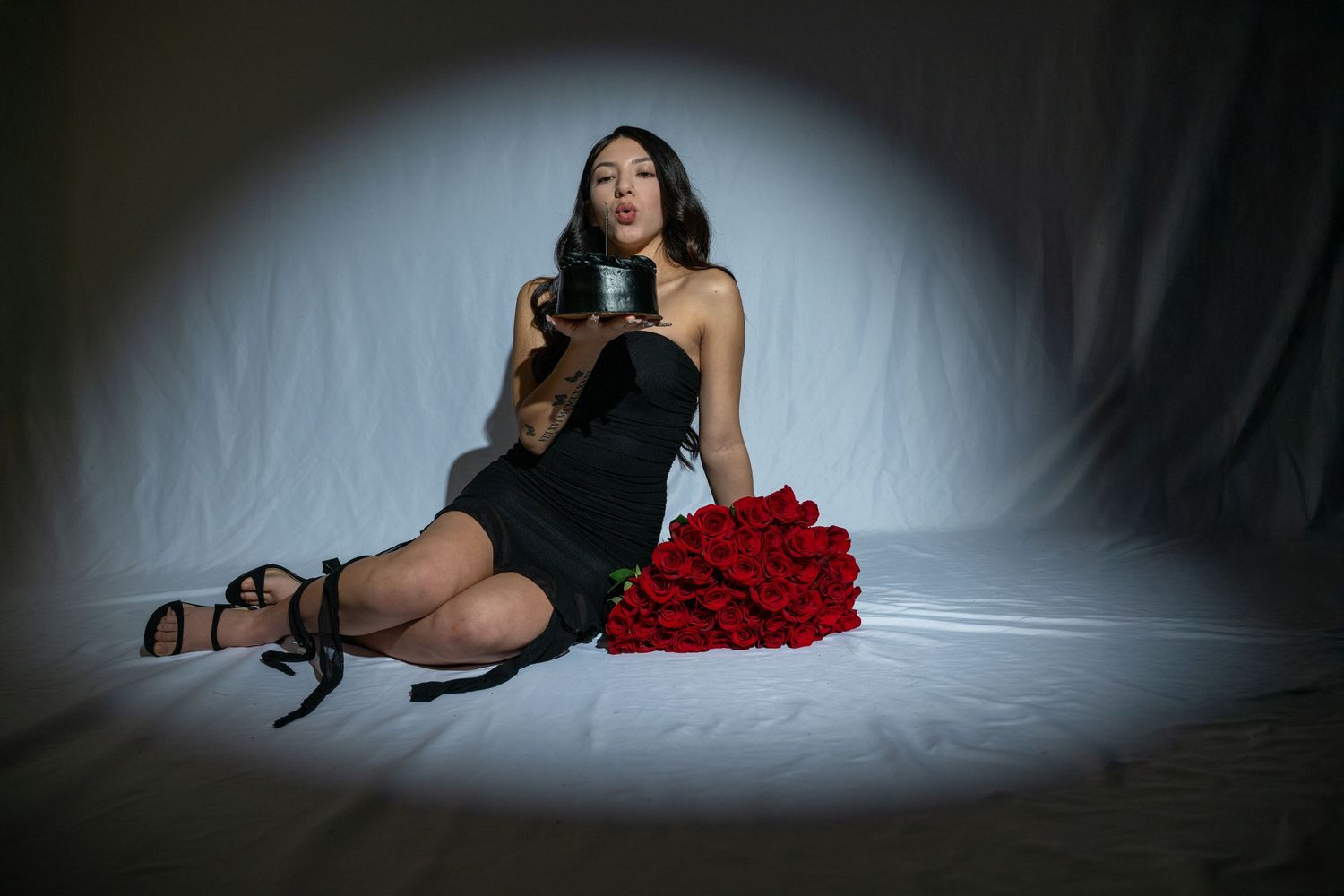 Woman in black dress sitting on floor holding a candle beside a bouquet of red roses.