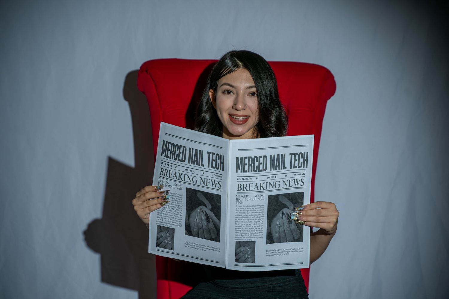 Woman smiling while reading Merced Nail Tech breaking news newspaper seated in red chair.