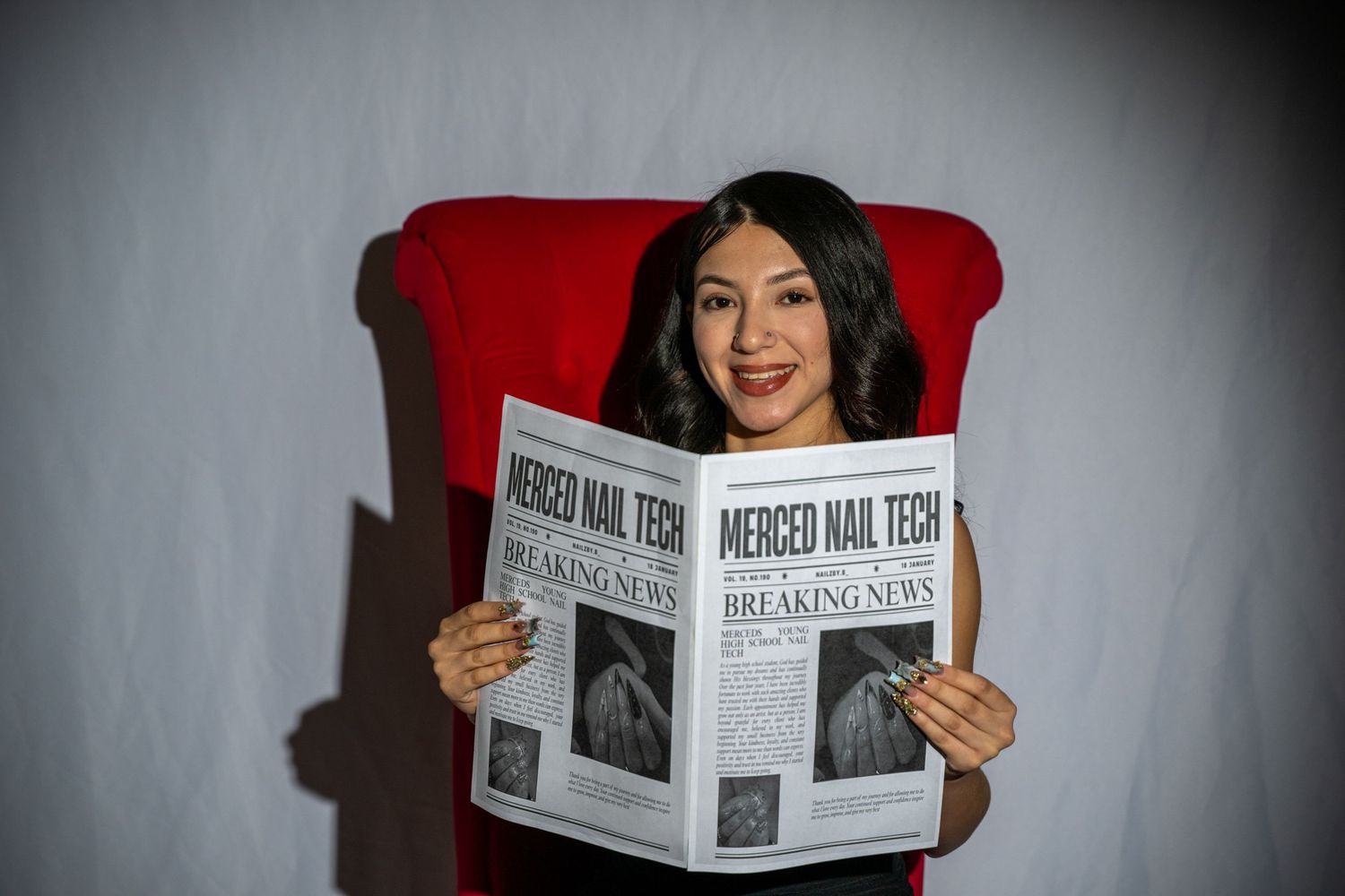 Woman sitting in red chair reading Merced Nail Tech Breaking News newspaper, smiling at camera.