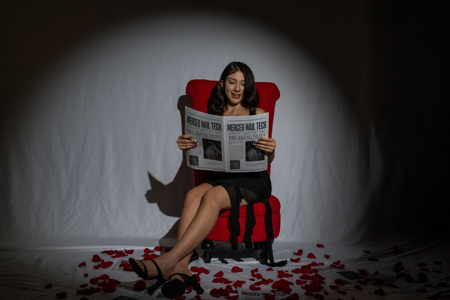 Woman reads newspaper in red chair surrounded by scattered rose petals on grey background.