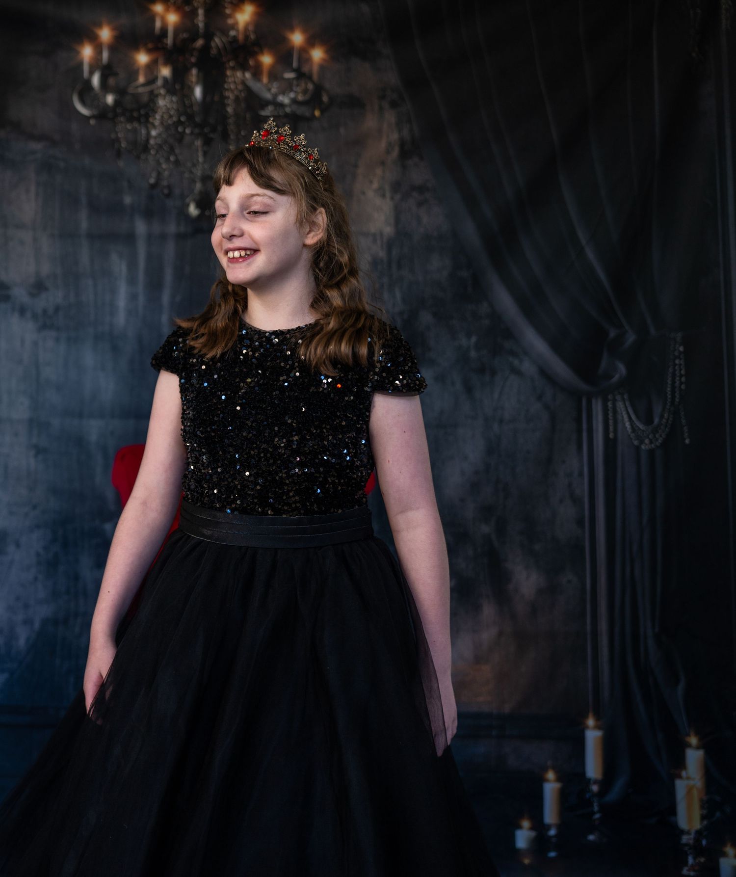 Person wearing sparkly black dress poses in dramatic studio setting with chandelier.