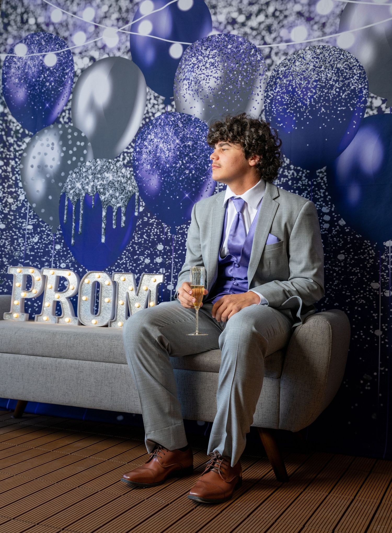 A student in a gray suit and purple tie sits alone on a couch at prom with dark blue and silver balloon decorations.