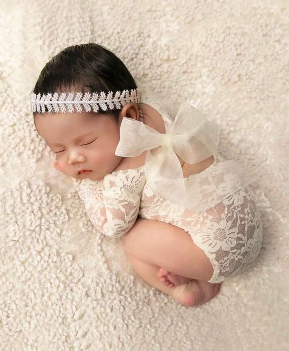 Baby in delicate white lace outfit resting on soft white textured fabric.