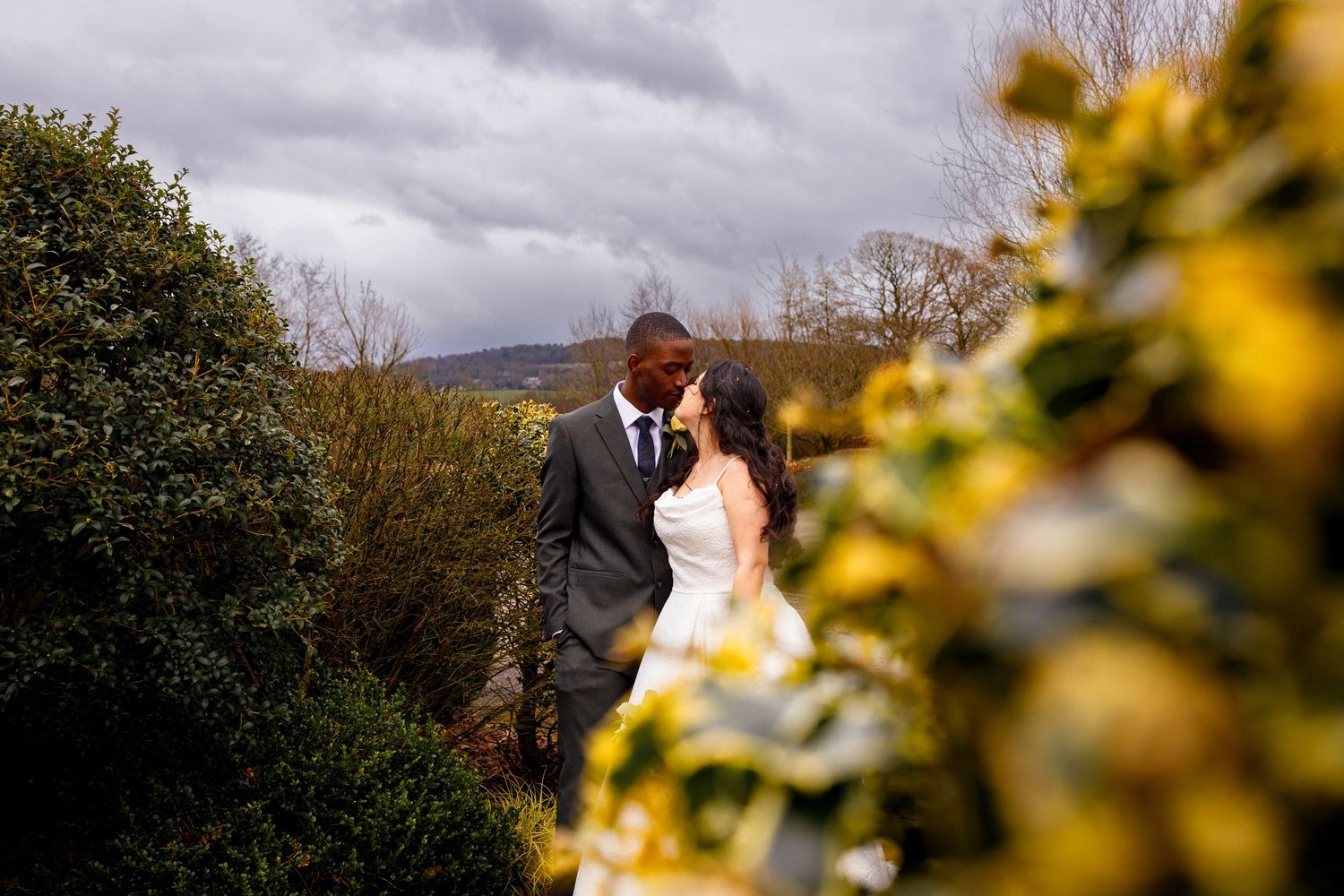 A romantic wedding couple embraces outdoors with yellow flowers in the foreground and moody storm clouds overhead.