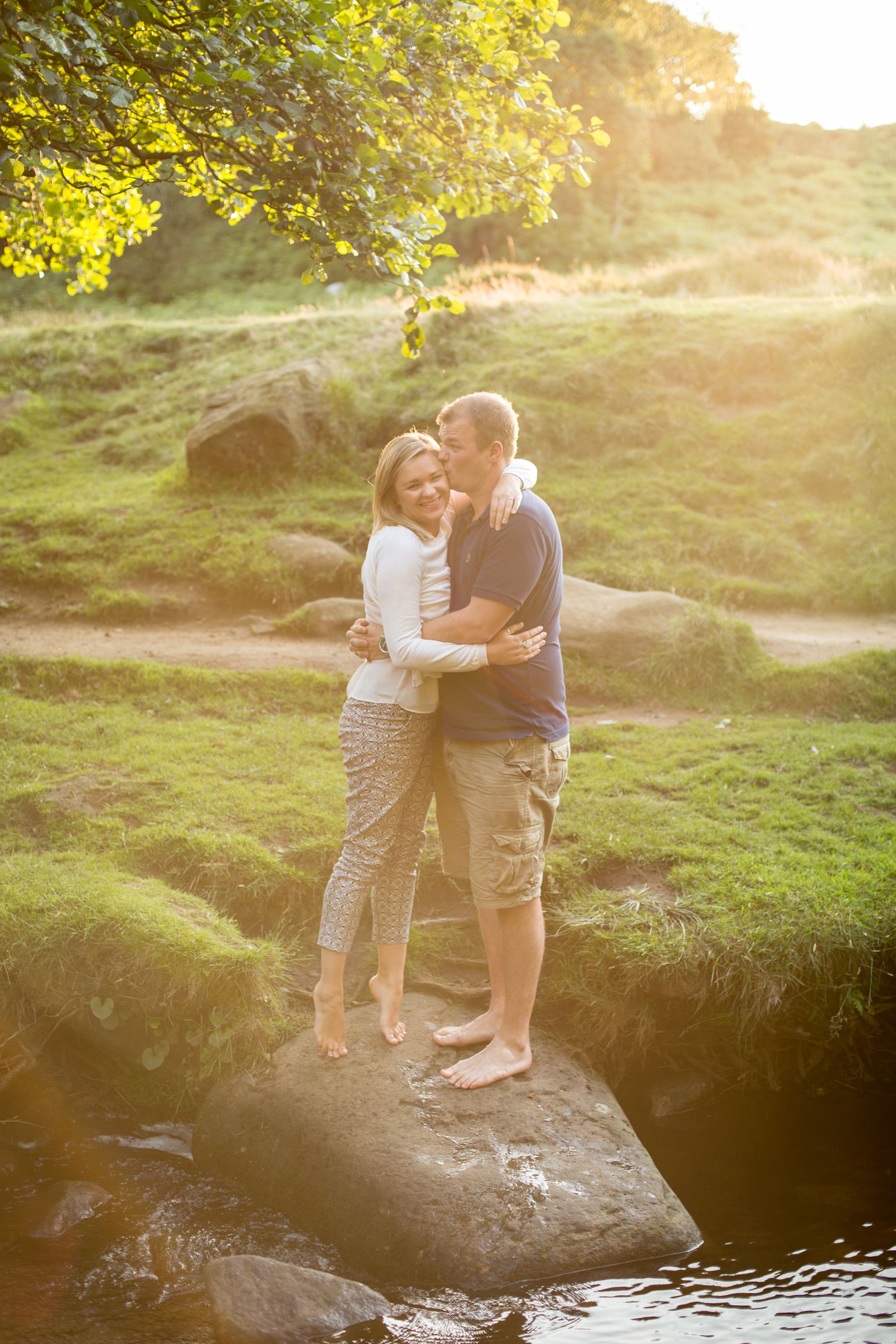 A couple embraces on a large rock by a stream during golden hour with sunlit foliage in the background.